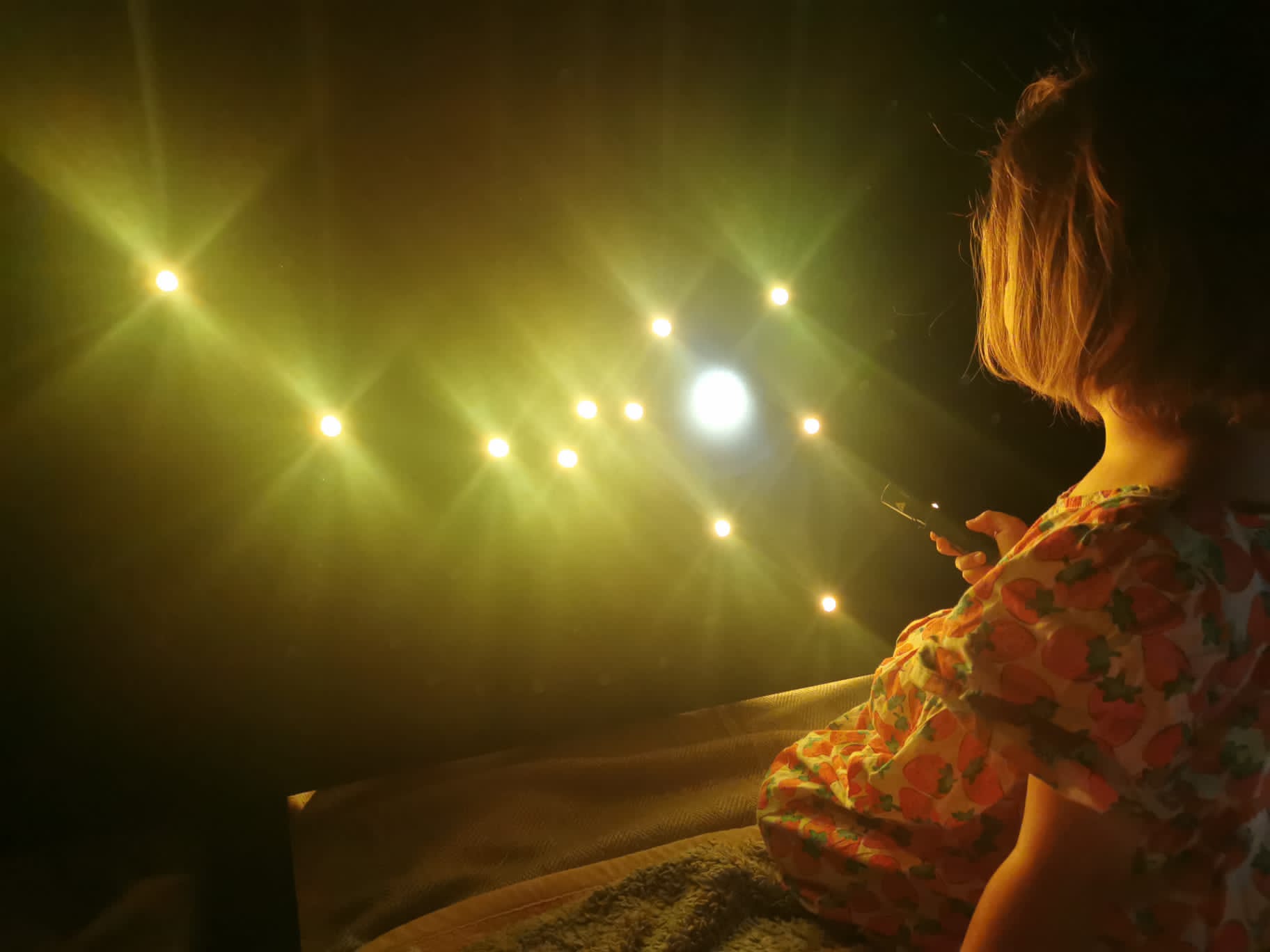  Leuchtende LEDs bilden Sternenbilder auf einer dunklen Fläche; ein Mädchen sitzt davor und richtet eine Taschenlampe auf die Installation.