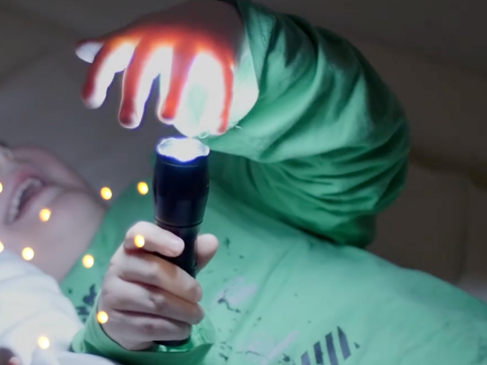 Ein Kind hält eine eingeschaltete Taschenlampe, deren Licht durch die andere Hand scheint, im Hintergrund sind warme Lichter im Innenraum zu sehen. 